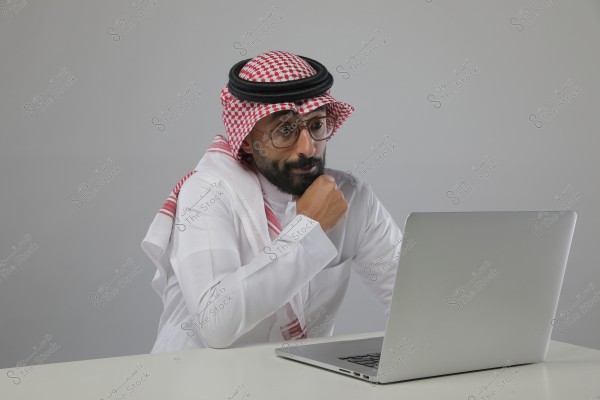 A portrait of a man sitting in front of a laptop. He is wearing traditional white attire and a red-checkered headscarf with a black rope band, suggesting Saudi origin. The man appears to be thoughtful or focused on the laptop screen. The background is grey, highlighting the man and the device.