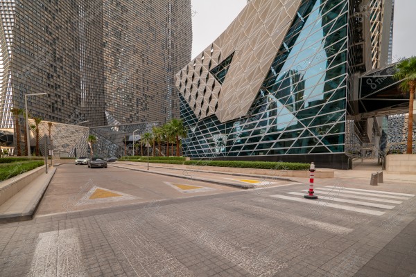 Image showing modern architectural buildings in an urban setting. The glass facade and angular structures are prominent features of the architecture, with trees and greenery lining the road. Two cars are parked on the road leading to the complex.