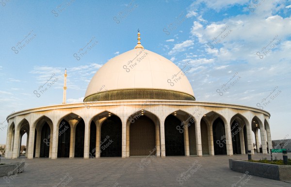 Image of a large mosque featuring traditional Islamic architectural design. The mosque has a prominent white dome and a single tall minaret, surrounded by a series of symmetrical arches that form the main entrances. The sky is blue with light clouds in the background, adding a picturesque touch to the scene.