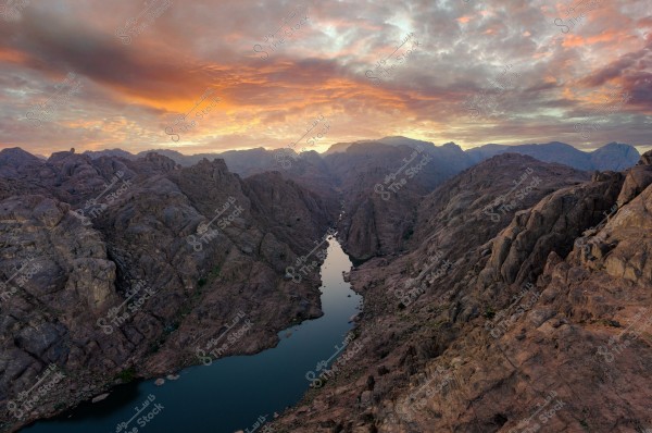 A natural landscape depicting a narrow valley between rocky mountains, with water reflecting in the river under the light of the sunrise. The sky is covered with orange-pink clouds, adding beauty to the natural scene.