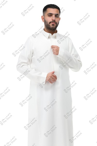 Image of a man wearing a long-sleeved white thobe, standing in front of a white background. The thobe has a traditional style, suggesting it might be from Saudi Arabia or the Gulf region. The man appears to be adjusting the collar of the thobe.