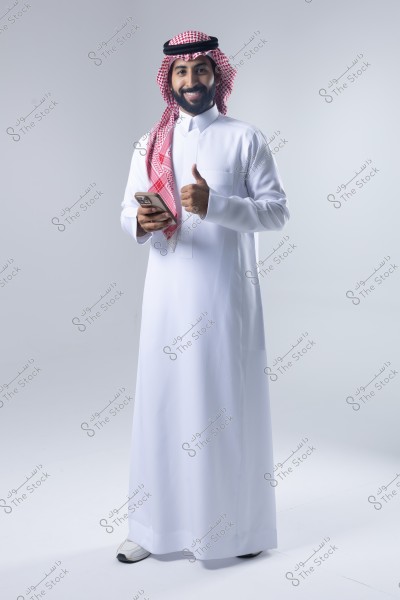 A portrait of an Arab man, wearing traditional Saudi attire, including a white thobe and a red ghutra with a black agal. He is holding a mobile phone in his right hand and giving a thumbs-up with his left hand, standing against a simple white background.