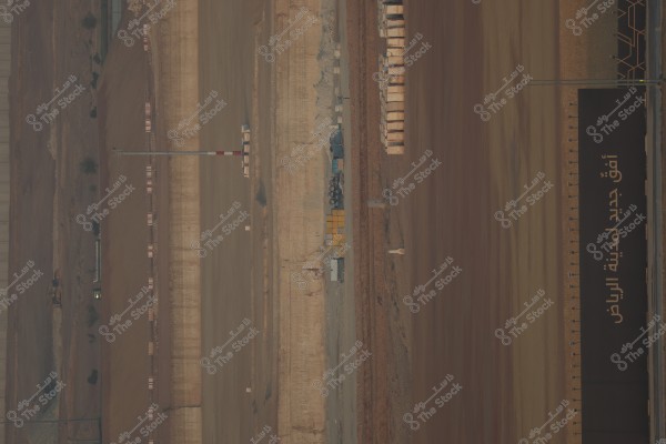 The image shows an aerial view of a large construction site near a desert area. On the right side of the image, there is a large black sign with Arabic writing indicating a new project in Riyadh. Two long dirt tracks run vertically through the center, flanked by soil and barriers. Some vehicles and machinery can be seen near the center of the track, alongside piles of construction materials.
