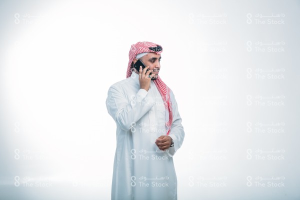 بورتريه رجل سعودي ، يرتدي الثوب ويقف أمام خلفية بيضاء، مبتسم ، يتحدث عبر الجوال ، مكالمات الأهل والأصدقاء ، تعابير وجه تدل على السعادة ، حوار ممتع مع شخص ما ، صورة شخصية