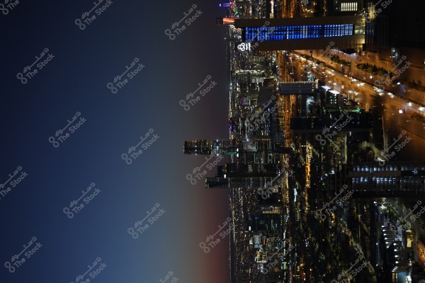 A nighttime cityscape illuminated by skyscraper lights and streetlights. Tall buildings stand out against the dark sky, with one prominently featuring blue lighting. Traffic motion is visible through car headlights and taillights on the streets.