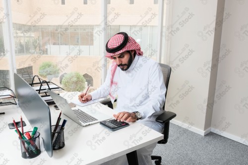 رجل سعودي يرتدي الزي السعودي التقليدي يعمل في مكتب بواجهة زجاجية ويستخدم قلم رصاص لتدوين الملاحظات