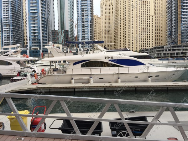 The image shows a white yacht with the number DT1459 docked at Dubai Marina. Surrounding the yacht are tall buildings and a metal railing in the foreground. Two other yachts are visible in the background with modern skyscrapers creating the backdrop.