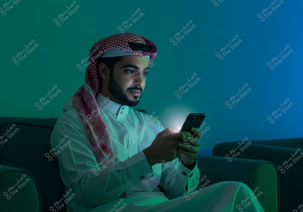 A portrait of a man sitting on a couch, wearing a white thobe and a red-checkered ghutra, holding a phone emitting a glowing light. He appears to be in a calm environment with dim blue and green lighting.