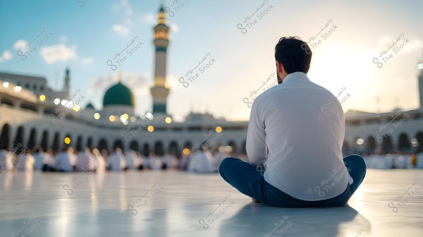 The image shows a person sitting cross-legged in the courtyard of Al-Masjid an-Nabawi in Medina, Saudi Arabia. The person is wearing a white shirt and blue pants, facing the mosque, which features the iconic green dome and prominent minaret in the background. The lighting suggests sunset, with soft lights shimmering in the square.
