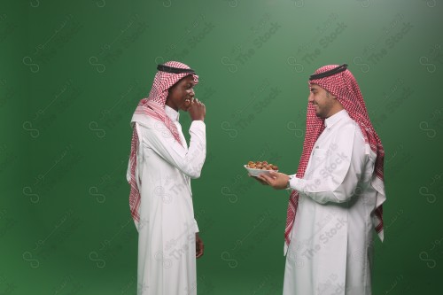 رجل سعودي يرتدي الثوب و الشماغ يقوم بتقديم الحلوى لرجل سعودي اخر