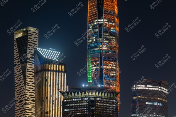 The image shows a group of skyscrapers illuminated at night in a modern city. The tower in the center is striking with orange and blue lighting, while other buildings exhibit various geometric designs. The dark sky enhances the prominence of the architectural lighting.