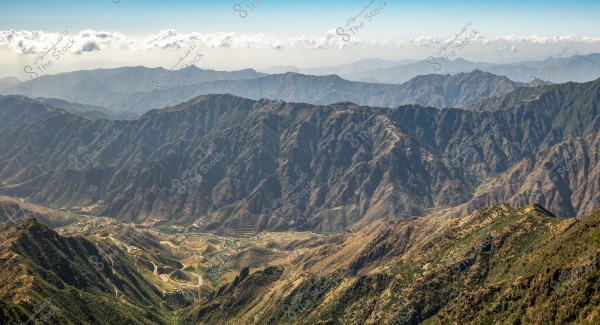An image of a mountainous landscape extending across a wide scene, with a range of mountains visible on the horizon. Multiple layered hills are draped in shadows with patches of greenery in some areas. Below, winding paths or roads are visible, showing the rugged terrain. The sky is clear with a few light clouds in the distance.