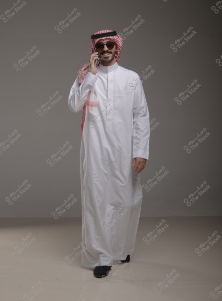 A portrait of a man wearing a traditional white thobe with a red shemagh and agal, talking on a mobile phone. He is wearing sunglasses and smiling. The setting appears to be a studio environment.