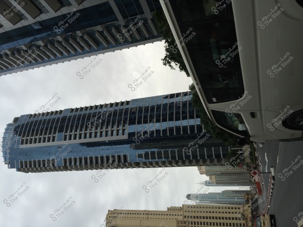 An image of an urban street in a modern city, featuring tall glass skyscrapers highlighting contemporary architecture. In the foreground, there is a white vehicle parked near the corner of the street. The sky is overcast, reflecting off the buildings in the background.