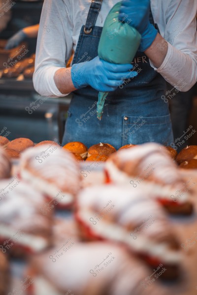 A baker wearing an apron and blue gloves fills pastries using a piping bag in a bakery.