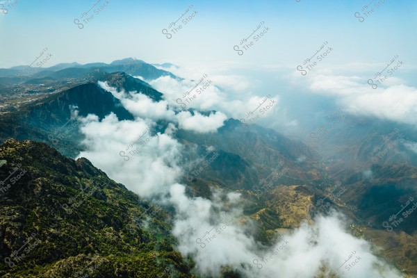 Aerial view of mountains partially covered by a thick layer of white clouds, stretching across the horizon in a stunning landscape. The mountainous terrain is dotted with sparse green vegetation, with details of a distant valley running between the mountain range.