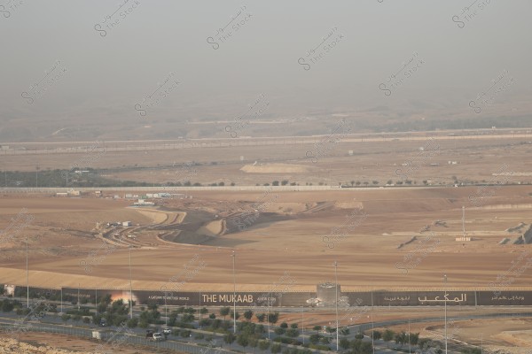 The image shows a landscape view of a construction site in a desert area. A wide flat land can be seen under a hazy sky, with dirt roads extending across the site and a few scattered trees. In the foreground, a large sign reads \"The Mukaab: A New Icon\" in both Arabic and English, indicating a new construction project.