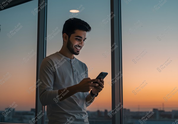 An image of a man standing by a large window overlooking a sunset view. The man is wearing a long-sleeved shirt and appears to be smiling while using a mobile phone. The sky is colored with shades of orange, pink, and blue, with buildings visible in the background.