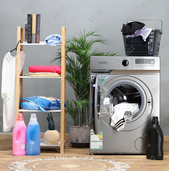 An image of a laundry space featuring an open silver washing machine with a black basket of clothes on top. A green plant in a pot is placed on the floor next to the washing machine. On the left is a wooden shelf with folded clothes and two books, and a hanger with a white shirt. At the bottom, there are bottles of laundry detergent in pink, blue, and black, alongside a large bag of laundry powder.