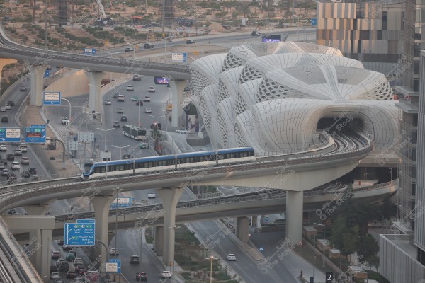 An image of a modern infrastructure in a city featuring an elevated metro train line adjacent to a uniquely designed building resembling marine creatures. Traffic is visible on the lower bridges and roads with numerous cars and buses. The surrounding buildings highlight the urban modernity of the scene.