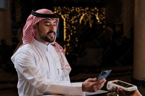 شاب سعودي في استقبال الفندق ، ويقوم بتعابير مختلفة باستخدام الجوال  ، ويقوم بدفع عبر جهاز الدفع الالكتروني