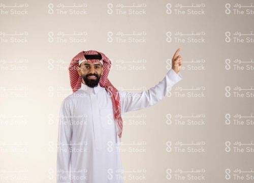 شاب سعودي مبتسم يرتدي الزي السعودي التقليدي يشير بإصبعه السبابة يسار الصورة، رجل سعودي ينقر على أيقونة افتراضية