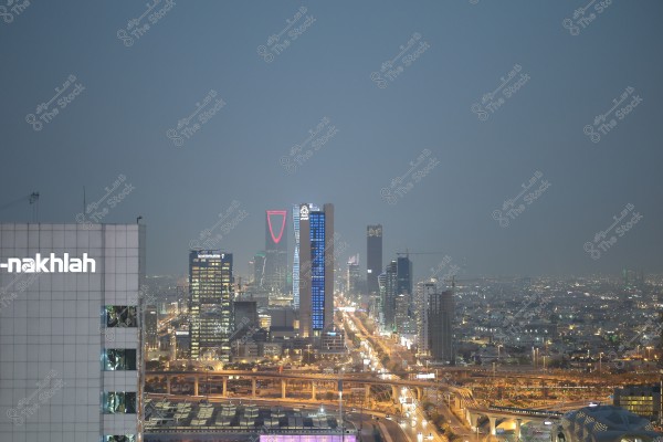An aerial view of Riyadh city in the evening, featuring a cluster of high-rise buildings under a soft blue sky. At the center, the Kingdom Tower is shown with its distinctive red lighting, while illuminated billboards can be seen on the nearby towers. Streets are lit by car headlights and street lamps, giving the city a lively and vibrant nighttime appearance.