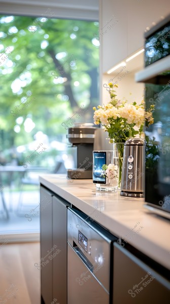 An image of a modern kitchen showing a countertop with a coffee machine, a glass vase containing white and cream-colored flowers, and a silver toaster. The window in the background reveals a green backdrop with trees and plants, suggesting a garden setting, and there is also a white dishwasher with a digital screen.
