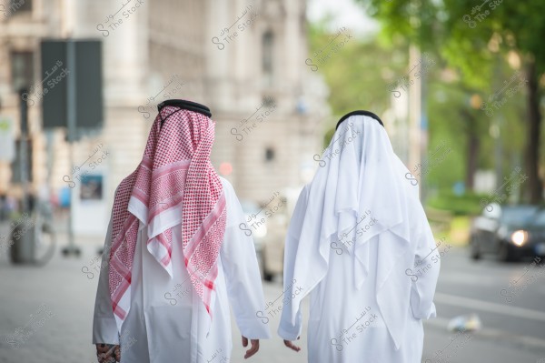 Image of two men walking on a street. The first man is wearing a white thobe and a red and white checkered ghutra with a black agal. The second man is wearing a white thobe and a plain white ghutra with a black agal. The background shows buildings and a road, with some trees nearby.