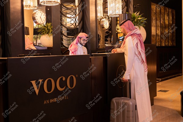 The image shows two men in a luxurious hotel lobby. Both men are wearing traditional Saudi attire, the thobe with the shemagh. One man is standing at the counter holding a suitcase, while the other is behind the counter smiling, seemingly during a check-in process. The background features plush decor including a wall clock, plants, and elegant lighting. The hotel name \"voco\" is displayed on the counter.