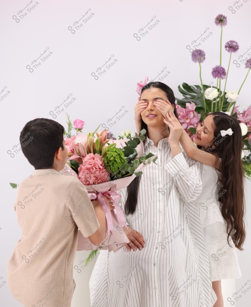An image of a woman in a long, striped white and black dress with a young girl in a white dress playfully covering the woman\'s eyes with her hands. There are various colorful flowers in the background, and a boy is holding a large bouquet of pink and green flowers in the foreground. The atmosphere appears festive and joyful.