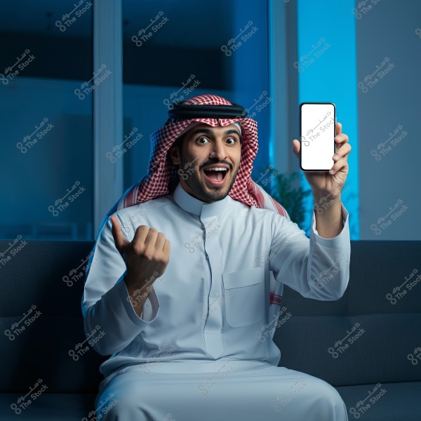 A photo of a man sitting on a couch wearing a white thobe and a red checkered headscarf. He looks happy, smiling broadly, and holding up a smartphone in his right hand with a bright white screen. The surrounding environment is dimly lit with a soft blue hue.