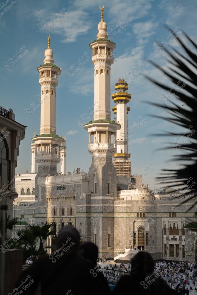 The image shows an exterior view of the Grand Mosque in Mecca, Saudi Arabia, highlighting the towering minarets and the grand architectural gates. The image also depicts crowds gathered for prayer or performing Umrah, with a clear sky and some trees visible in the background.