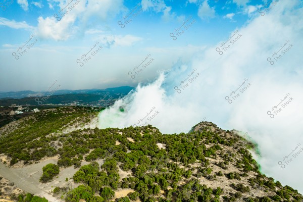 An aerial view of a mountainous area covered with green vegetation under a beautiful blue sky. Thick mist rolls over the slopes, creating a mystical atmosphere in the scene.