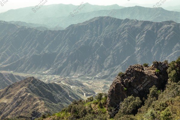 A mountain landscape showing a range of rugged peaks stretching across the horizon. In the foreground, there are green hills with scattered plants and trees, while the background features larger mountains covered in shadows, with agricultural areas in the valley below.