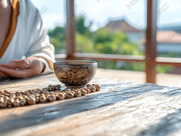 An image showing a person\'s hand wearing white clothing with brown edges, resting on an old wooden table. On the table, there is a small brown bowl and a prayer bead string made of natural beads. In the background, there is a view of a natural landscape seen through a window.