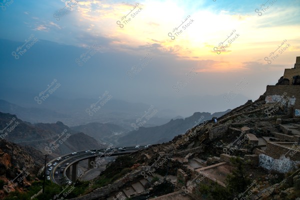 Stunning mountain view featuring a sunset on the horizon with a sky in gradient shades of blue and orange. Below, a road winds through the mountains, rising towards the sky, with traffic flowing on it. On the right side, remnants of stone buildings and rocky terrain are visible, with a high bridge in the middle. The background mountain view is hazy and expansive.