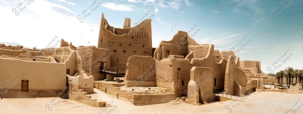 A view of a heritage village consisting of traditional mud buildings showcasing old Najdi architecture. The image features clay walls with small windows and wooden doors, adorned with distinctive geometric patterns on some walls. In the background, a blue sky and several palm trees are visible, reflecting the character of the Arabian desert environment.