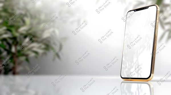A modern smartphone with a gold frame positioned on a glossy white surface. The screen is reflective with no visible content. The background features blurred green plants.