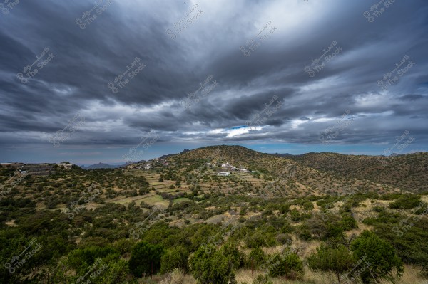 An image of a natural countryside landscape featuring hills covered with green trees and shrubs under a densely cloudy sky. Small houses are scattered across the hills, appearing as part of a rural village amidst the peaceful nature.