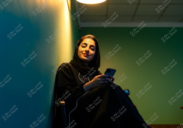 An image of a woman sitting on a chair next to a light green wall. She is wearing a black abaya and a black headscarf. She is holding a mobile phone and appears relaxed and smiling. A ceiling light casts a warm glow over the scene.