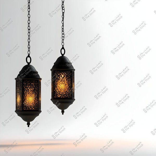 Two ornate hanging lanterns with intricate patterns hang on metal chains. The internal lighting gives a warm glow to the lanterns. The background is soft and white, highlighting the detailed design of the lanterns.
