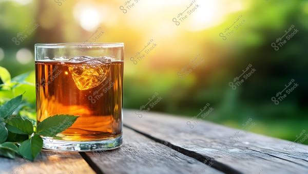 Image of a glass filled with a brown beverage and ice cubes, placed on a wooden table in a garden. There are some green leaves next to the glass, and the blurred background shows trees and plants with the warm glow of the sunset.