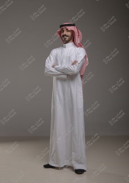 صورة لرجل يرتدي الثوب التقليدي السعودي الأبيض والغترة الحمراء, واقفًا بذراعين متقاطعتين على خلفية رمادية.