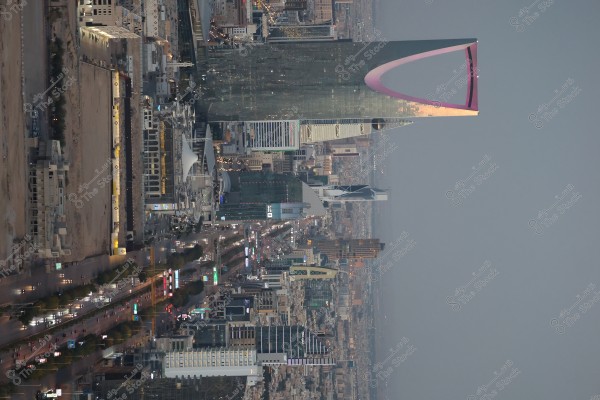 An aerial view of Riyadh, Saudi Arabia, showcasing the main road and towering buildings, including the distinctive Kingdom Tower. The street is bustling with lights and vehicles, surrounded by various buildings.