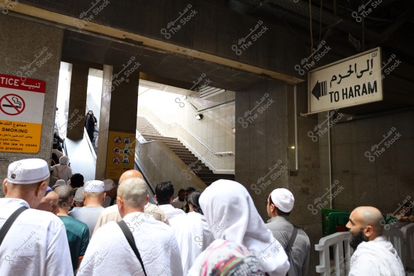 Image of a group of people wearing Ihram clothing or traditional garments, preparing to ascend an escalator. A large sign points the direction \"To Haram,\" and another sign prohibits smoking. The location appears to be an entrance or corridor with dim lighting and marble walls.