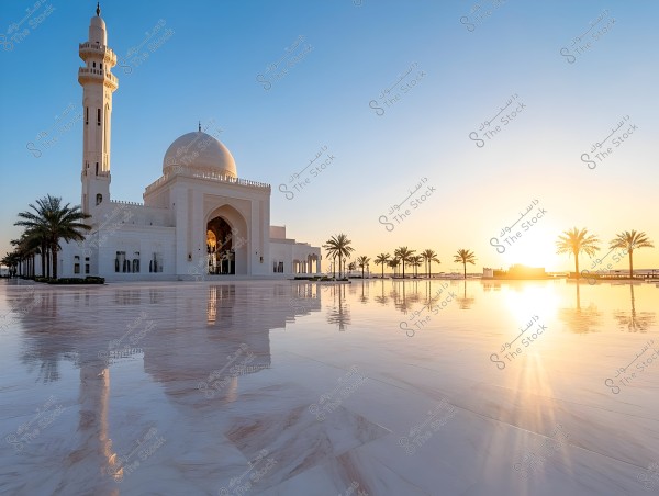 An image of a large mosque with white pillars and a grand dome, featuring a minaret on the left side. The mosque is surrounded by palm trees, whose reflections are visible on a glossy marble floor. The sun is rising or setting in the background, casting a warm light over the scene.