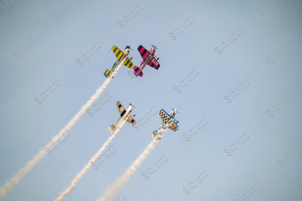 Four colorful airplanes performing an aerial display in the clear blue sky. The airplanes are releasing white smoke as they fly in a slanted formation. The topmost plane is colored black and yellow with blue stripes, the middle plane is pink and black, another one is blue and black with checkered details, and the fourth one is light gray.