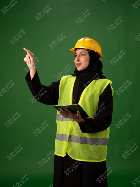 Image of a woman wearing a yellow construction helmet and a light green reflective safety vest, holding a tablet and pointing with her finger. She is wearing a black abaya and is set against a green background.