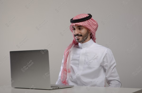 صورة لرجل يجلس خلف جهاز كمبيوتر محمول, يرتدي الزي السعودي التقليدي الذي يتضمن الثوب الأبيض والغترة الحمراء والعقال الأسود. يبدو الرجل مبتسمًا وينظر إلى شاشة الكمبيوتر. الخلفية بسيطة وحيادية.